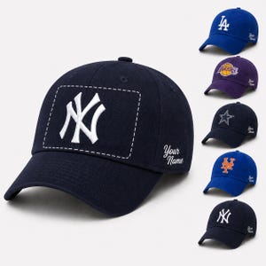 May include: A collection of baseball caps in various colours including navy, royal blue, and purple. Each cap features a sports team logo on the front, a stitched rectangular border, and the embroidered text Your Name on the side panel.