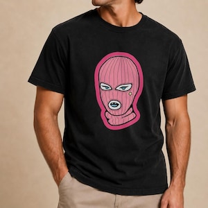 May include: A black short-sleeved t-shirt featuring a graphic print of a pink balaclava mask. The mask design includes stylized eyes, a small yellow star near the eye, and a blue mouth opening, all outlined in a thick pink border.