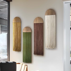 Puede incluir: Cuatro tapices de pared con una parte superior de madera y largos hilos de lana en diferentes colores. Los colores son amarillo mostaza, verde, marrón y blanco.