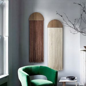 Puede incluir: Dos tapices de macramé de pared con arcos de madera, uno marrón y otro blanco, colgados en una pared gris claro sobre un sillón de terciopelo verde y una pequeña mesa auxiliar de madera.