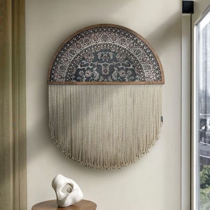 Pode incluir: Uma grande tapeçaria de parede semicircular com um tecido estampado floral escuro na parte superior e longas franjas creme. A moldura de madeira é castanho quente. Uma pequena mesa lateral de madeira e uma escultura branca estão em primeiro plano.