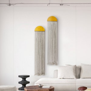 Puede incluir: Dos tapices de macramé blancos con acentos amarillos en forma de semicírculo en una sala de estar moderna con un sofá blanco, una mesa de café de madera y una mesa auxiliar negra.