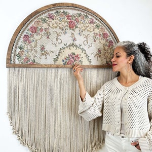 May include: A large, round wall hanging with a floral pattern framed in wood. The hanging has a fringe of white yarn. A person is standing next to the hanging, touching the fringe.