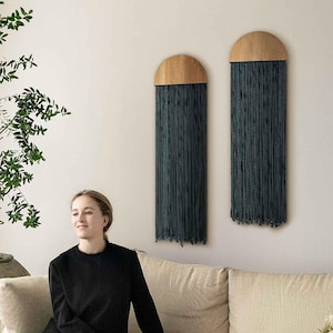 May include: A woman in black sits on a cream sofa. Two wall decorations with wooden tops and dark blue fringe are on the wall. A large potted plant and a coffee table with flowers are also in the room.