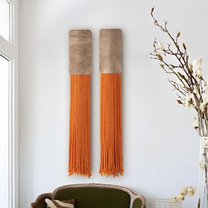 Może przedstawiać: Dwa wiszące ozdoby ścienne z drewnianym ramą i pomarańczowymi frędzlami z włóczki. Ozdoby wiszą na białej ścianie z zieloną welurową sofą i wazą z białymi kwiatami w tle.