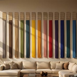 Può includere: Un soggiorno moderno con un divano componibile beige, un tavolino in legno e arazzi colorati. L'arte murale è composta da fili di tessuto verticali in vari colori, tra cui grigio, marrone, verde, giallo e blu. Sculture e un tavolino completano l'arredamento.