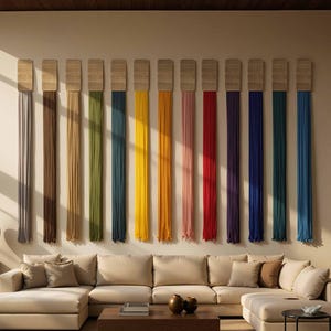 Può includere: Un grande divano angolare beige dal design basso, posizionato davanti a una parete con dodici arazzi verticali. Ogni decorazione è composta da una parte superiore rettangolare in legno con lunghe nappe in tessuto di vari colori.