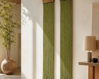 May include: Two olive green wall hangings with wooden tops and long, dangling strands. The hangings are displayed on a white wall, adding a touch of natural texture and color to the space. The design is simple and elegant.