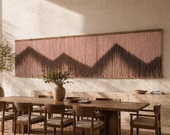 Puede incluir: Un comedor con una gran mesa de madera y sillas, una alfombra tejida y un tapiz de pared con un diseño geométrico rosa y marrón. Dos grandes jarrones con plantas flanquean la mesa. La habitación tiene una paleta de colores neutros.