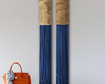 May include: Two wall hangings with wooden tops and long, blue fringe. An orange handbag and a scarf rest on a gray tufted bench with metal legs. The wall is painted a light grey.