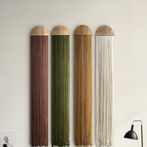 Könnte beinhalten: Vier vertikale Wandbehänge mit Holzaufsätzen und Strängen in Braun, Grün, Gold und Weiß. Ein dunkelbraunes Ledersofa und eine schwarze Lampe befinden sich im Vordergrund. Die Wandbehänge verleihen dem Raum Textur und Farbe.