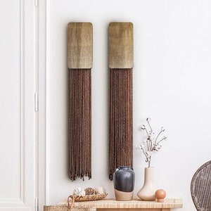 Op de afbeelding: Twee bruine en gouden wandhangers met lange kwastjes die aan de onderkant hangen. De hangers hangen aan een witte muur in een kamer met een houten bank, een stoel en een vaas met katoenen stelen.