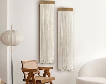 May include: Two white macrame wall hangings with wooden dowels, a white ottoman, a wooden chair, and a white sofa in a modern living room.