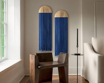 Op de afbeelding: Twee blauwe en gouden macramé wandhangers met een afgeronde bovenkant, hangend aan een witte muur. Een donkere houten stoel met een gebogen rugleuning en een donkere houten bijzettafel met een zwarte kaarsenhouder en een zwarte ronde basis staan op de voorgrond.