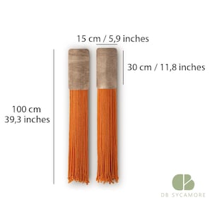 May include: Two orange tassel wall hangings with wooden tops. Each tassel is 100 cm long and 30 cm wide. The wooden tops are 15 cm wide.