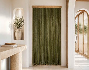 May include: Olive green beaded curtain hanging in a doorway. The curtain is made of numerous vertical strands, creating a textured, layered effect. A wooden header and a light-colored wall frame the curtain. The room has a neutral color palette.