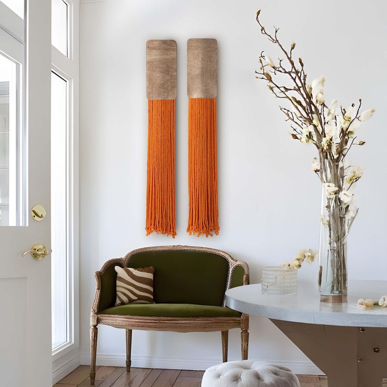 May include: Two wall hangings with a wood frame and orange yarn fringe. The hangings are on a white wall with a green velvet sofa and a vase of white flowers in the background.