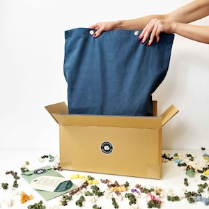 May include: A blue fabric bag is being pulled out of a cardboard box. The box has a sticker with the text "Crayola" on it. The bag is being pulled out by a hand. There are colourful yarn scraps scattered around the box.