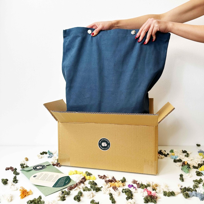 May include: A blue fabric bag is being pulled out of a cardboard box. The box has a sticker with the text "Crayola" on it. The bag is being pulled out by a hand. There are colourful yarn scraps scattered around the box.