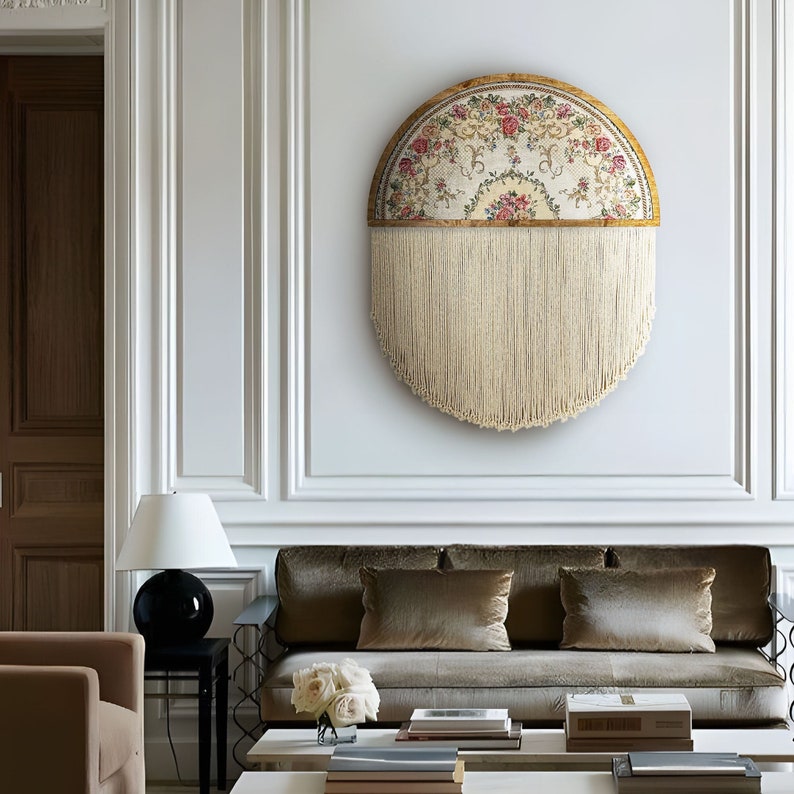 May include: A round wall hanging with a floral tapestry design and a fringe border. The tapestry is framed in wood and has a cream background with pink and red flowers. The fringe is a light cream colour.