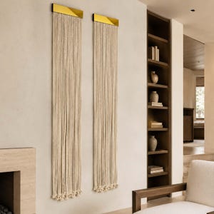 May include: Two wall hangings featuring a gold metal top and long, cream-coloured rope strands. The rope strands hang down, creating a textured, vertical design. The wall hangings are mounted on a light-coloured wall, adding a touch of elegance to the space.