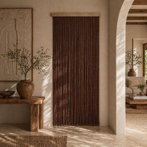 Op de afbeelding: Een donkerbruin kralengordijn hangt in een deuropening, met een lichte houten kop. Het gordijn is gemaakt van vele verticale strengen. De kamer heeft een neutraal kleurenpalet, met een houten bank en een grote vaas met groen.