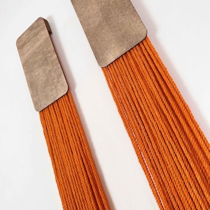 May include: Two wall hangings with a wooden rectangle at the top and a long fringe of orange yarn hanging down.