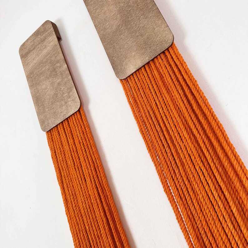 May include: Two wall hangings with a wooden rectangle at the top and a long fringe of orange yarn hanging down.