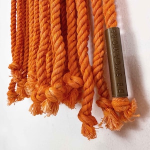 May include: Close-up of a bright orange macrame wall hanging with a brown wooden tag that reads "DB SYCAMORE".