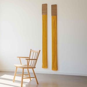 May include: Two wall hangings with wooden tops and long, yellow fringe. A light-colored wooden chair with a curved seat and armrests sits in front of a white wall. The wall hangings add a pop of color.