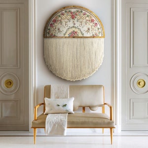 May include: A half-circle wall hanging with a floral tapestry design and a fringe of white yarn. The tapestry is framed in a wooden circle with a gold finish. The wall hanging is displayed on a white wall above a light beige sofa with a white throw pillow.