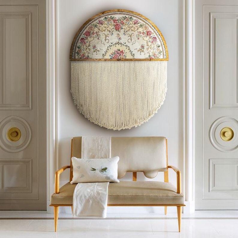 May include: A half-circle wall hanging with a floral tapestry design and a fringe of white yarn. The tapestry is framed in a wooden circle with a gold finish. The wall hanging is displayed on a white wall above a light beige sofa with a white throw pillow.