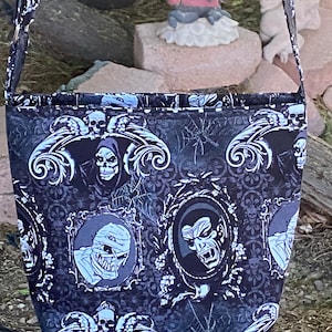 May include: Black and white Halloween-themed tote bag with a repeating pattern of skulls, monsters, and gothic designs. The bag has a black strap and is made of fabric.