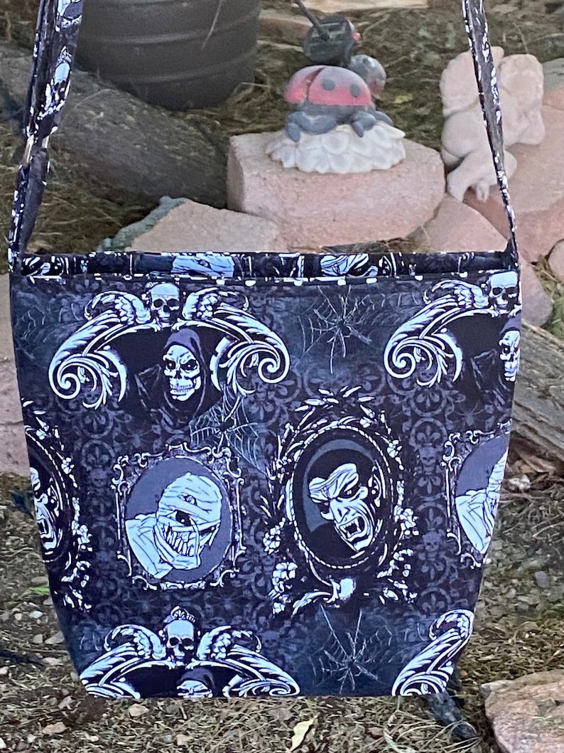 May include: Black and white Halloween-themed tote bag with a repeating pattern of skulls, monsters, and gothic designs. The bag has a black strap and is made of fabric.