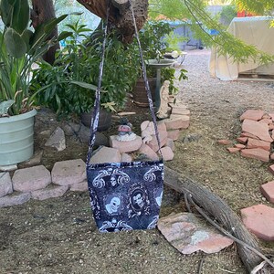 May include: A black and white fabric crossbody bag with a skull and floral pattern. The bag has a long strap and is hanging from a tree branch.