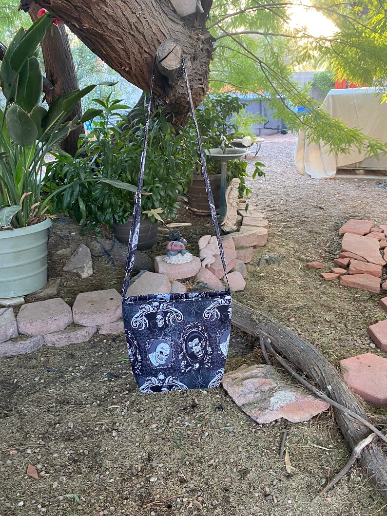 May include: A black and white fabric crossbody bag with a skull and floral pattern. The bag has a long strap and is hanging from a tree branch.