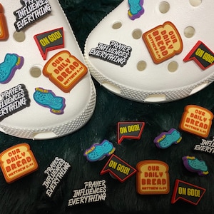 May include: A collection of colorful shoe charms with religious messages. The charms feature phrases like "Our Daily Bread", "Prayer Influences Everything", and "On God!" The charms are made of a soft, rubbery material and are designed to be attached to shoes.