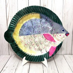 May include: A decorative ceramic plate in the shape of a fish. The plate features a green rim, a yellow and blue body, and pink fins. The fish-shaped plate is supported by a white stand.
