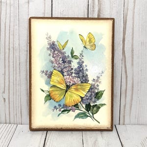 May include: A rectangular wooden plaque featuring a watercolor painting of yellow butterflies and purple lilac flowers. The butterflies have orange and yellow wings. The background is light blue and cream-colored. The plaque has a brown border.