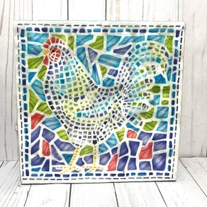 May include: A square mosaic artwork featuring a rooster. The rooster is depicted in white with a red comb and wattle, set against a background of blue, green, and red mosaic tiles. The artwork is framed with a white border.