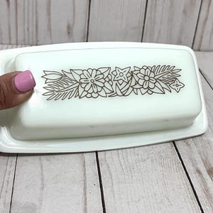 Könnte beinhalten: Eine weiße Butterdose mit rechteckigem Deckel und Boden. Der Deckel hat ein braunes Blumendesign. Die Dose steht auf einer hellen Holzoberfläche.
