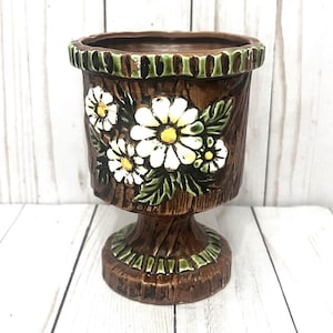 Puede incluir: Una maceta de cerámica marrón con base de pedestal, decorada con margaritas blancas y hojas verdes. El borde y la base tienen detalles verdes. La maceta tiene una apariencia texturizada similar a la corteza.