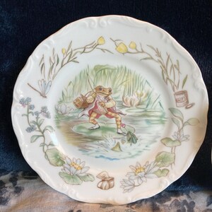 May include: White ceramic plate with a scalloped edge, depicting a frog fishing in a pond. The design includes water lilies, a dragonfly, and the word "BAIT" on a small container. Decorative floral border. The plate's diameter is approximately 20 cm.