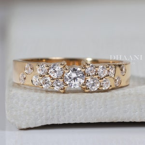 Puede incluir: Un anillo de banda de oro con un racimo de pequeños diamantes y un diamante central más grande. El anillo está colocado sobre una superficie blanca.
