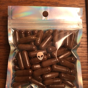 Può includere: Un sacchetto di plastica trasparente riempito con circa 50 capsule marroni. Il sacchetto ha una finitura olografica iridescente e un piccolo foro nella parte superiore.