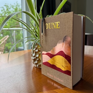 Puede incluir: Un libro de tapa dura titulado "DUNE" de Frank Herbert. El libro tiene una cubierta de lino marrón con letras doradas y un diseño de paisaje desértico colorido. Una cinta negra como marcapáginas es visible. El libro está sobre una mesa de madera.