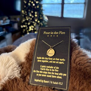 May include: A gold-colored necklace with a pendant featuring a triangle and three small stones, displayed on a black card. The card reads "Four in the Fire" and includes inspirational text. The necklace rests on a brown and cream-colored blanket.