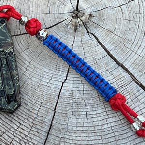 May include: A red and blue paracord keychain with silver beads and a knot at the end. The keychain is attached to a camouflage pocket knife.