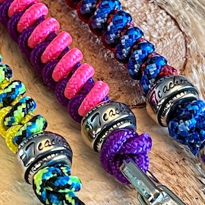 May include: Three colorful paracord bracelets with silver metal beads that say "Teacher". The bracelets are purple and pink, blue and red, and black and yellow.