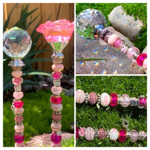 May include: Two pink and silver garden stakes with crystal tops and pink beads. The stakes are decorated with pink ribbon charms and pink and silver beads. The stakes are on a green grass background.